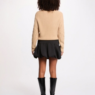 Mulher de costas com casaco de malha bege, saia preta bufante e botas pretas até ao joelho