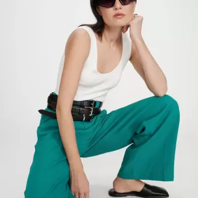 Mulher com óculos de sol, blusa branca, calças verdes largas, cinto preto e sapatos pretos num fundo branco