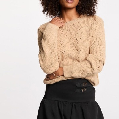 Mulher com camisola bege de malha e saia preta com fivelas em fundo branco