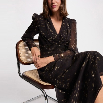 Vestido preto comprido com padrão dourado e mangas transparentes, mulher sentada numa cadeira palhinha e metal.