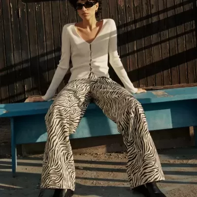 Mulher com sweater branco, calças de padrão animal preto e branco, botas pretas e óculos escuros sentada num banco azul