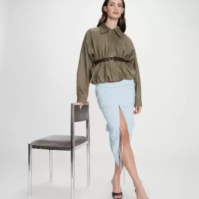 Modelo com camisa verde-oliva cinto castanho saia azul claro riscas salto castanho ao lado de cadeira metálica cinza