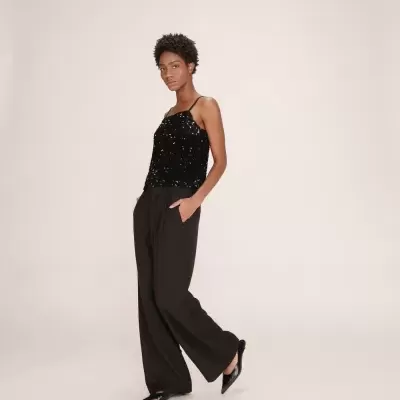 Mulher com top preto de lantejoulas e calças largas pretas num fundo neutro