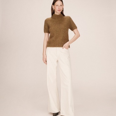 Mulher com camisola castanha de malha e calças brancas largas, fundo bege.