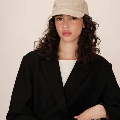 Mulher com casaco preto, camiseta branca e boné bege com texto branco no fundo neutro