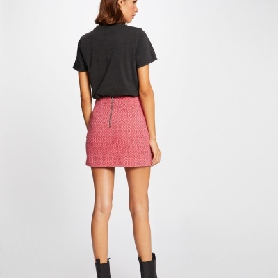 Modelo de costas usando saia curta rosa com padrão texturizado, t-shirt preta e botas pretas em fundo branco.