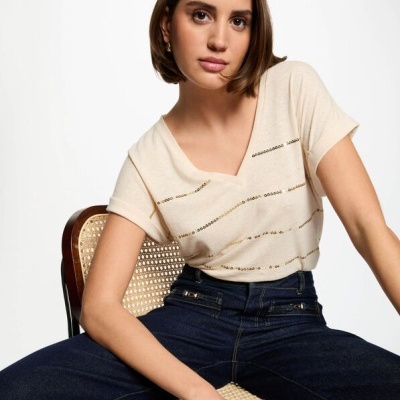 Mulher com camisola creme à riscas douradas e jeans escuros sentada numa cadeira de madeira com palhinha.