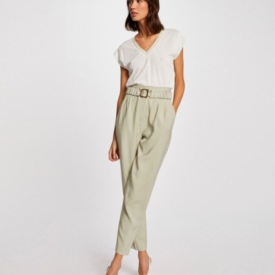Mulher com t-shirt branca de gola V, calças bege com cinto largo e sapatos brancos de salto, fundo branco.