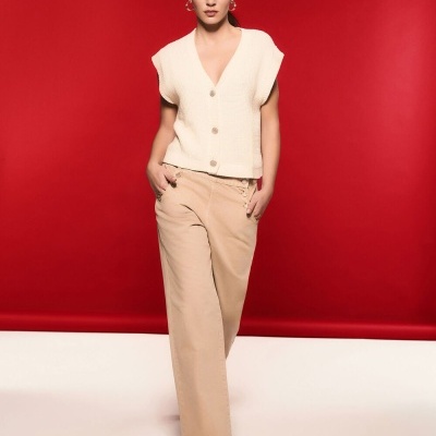 Mulher com colete creme de malha e calças largas bege num fundo vermelho