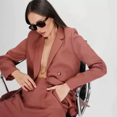 Mulher com fato rosa velho, óculos escuros, sentada numa cadeira de escritório com fundo branco.