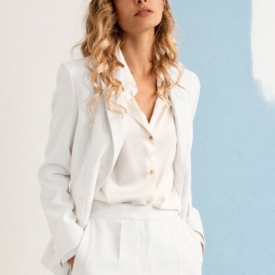 Mulher com fato branco composto por blazer, calças e camisa branca, em frente a parede branca com faixa azul