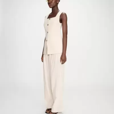 Pessoa com colete bege de botões, calças largas bege e sandálias pretas em fundo branco