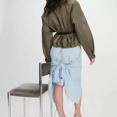 Mulher de costas com casaco verde oliva, saia azul claro amarrada e sapatos de salto perto de cadeira metálica.