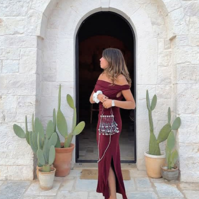 Mulher com vestido bordô longo e saco bordado à frente de porta arqueada de pedra com vasos de cactos.