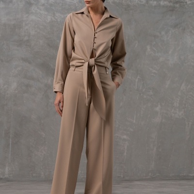 Mulher usando macacão bege com cinto e sapatos de salto animal print em fundo cinza.