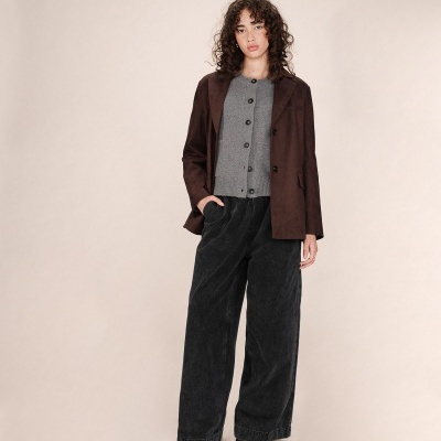 Mulher com blazer castanho, camisola cinza, calças largas pretas e sapatos pretos em fundo branco