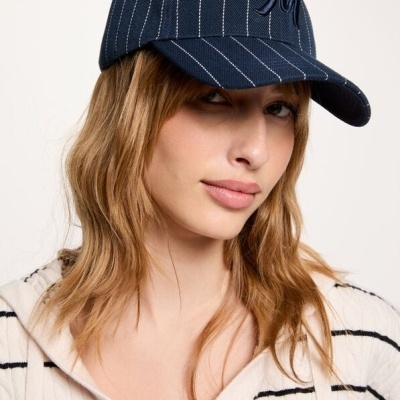 Boné azul escuro com riscas brancas e logótipo, usado por mulher com casaco riscado