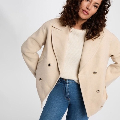 Casaco bege de duplo abotoamento com botões metálicos, usado por mulher com camisola creme e jeans azuis