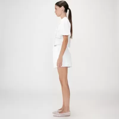 Mulher de perfil com conjunto branco com padrão de perfurações, blusa e calções, calçando sapatilhas nude.