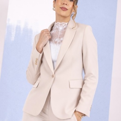 Conjunto feminino bege com blazer e calças, blusa branca de renda, fundo azul e rosa.