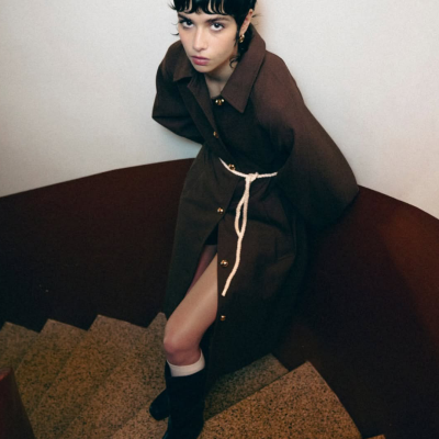 Mulher de casaco castanho escuro com cinto branco numa escada interior