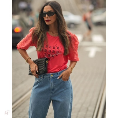 Mulher com t-shirt coral, calças de ganga, óculos de sol e carteira preta numa rua com carris de elétrico