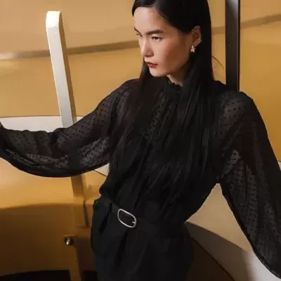 Blusa preta transparente de bolinhas com cinto preto em modelo junto a escadas interiores.