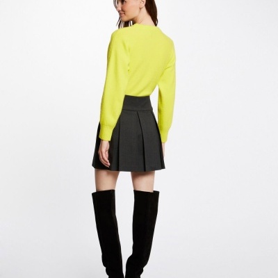 Mulher de costas com camisola amarela, saia preta plissada e botas pretas de salto em fundo branco