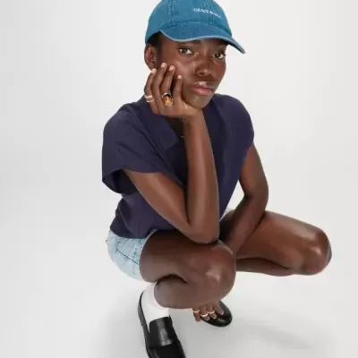 Pessoa com boné azul, camisa roxa, calções de ganga e sapatos negros agachada sobre fundo branco