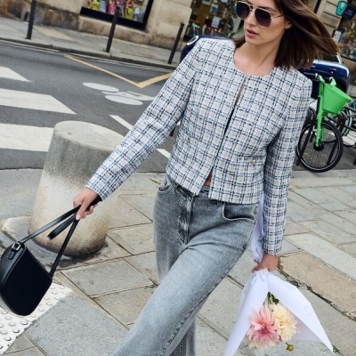 Mulher com casaco tweed azul e branco, jeans cinza, óculos escuros, mala preta e bouquet de flores numa rua urbana