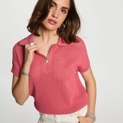 Camisola rosa de malha com gola e fecho, calças brancas e acessórios prateados.