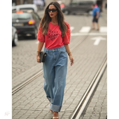 Mulher com camisa vermelha de mangas bufantes e calças de ganga azuis claras caminhando na rua com carris de elétrico