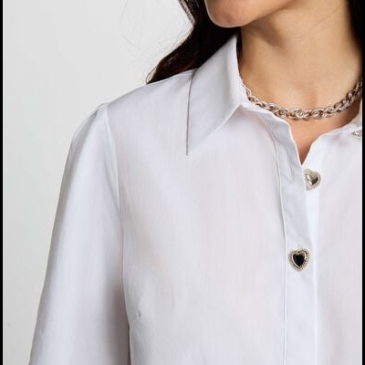 Blusa branca com botões de coração dourado e colar prateado num fundo cinza claro.