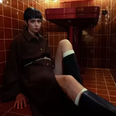 Mulher com sobretudo castanho e botas pretas sentada em casa de banho com azulejos castanhos.