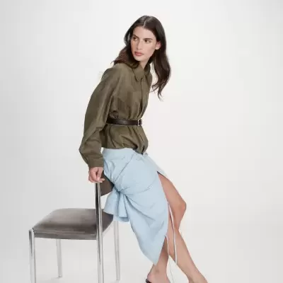 Mulher sentada numa cadeira vestindo blusa verde militar e saia azul clara com salto castanho.