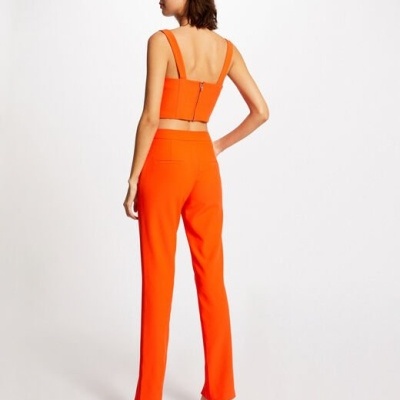 Mulher vista de costas com conjunto laranja composto por top cropped e calças, usando sandálias de salto alto laranja, em fundo branco.
