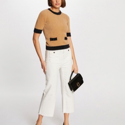 Mulher com camisola castanha e preta, calças brancas, sapatos pretos e mala preta diante fundo branco