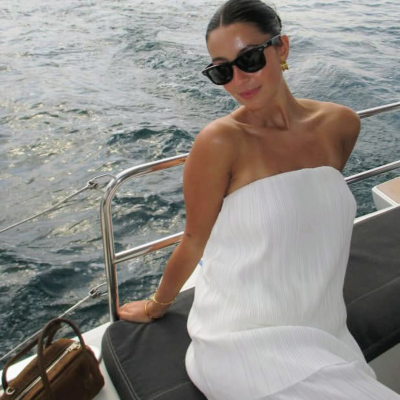 Mulher com vestido branco plissado, óculos escuros pretos, sentada num barco com mala castanha e mar ao fundo