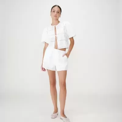 Mulher vestindo conjunto branco com blusa de manga curta e calções com padrão de ilhós, sapatos baixos transparentes, fundo branco.