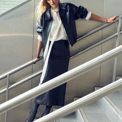 Mulher vestindo casaco de couro preto, blusa branca e saia preta numa escada.