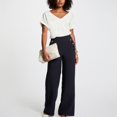 Mulher com camisola branca e calças pretas, segura mala cremosa em fundo branco