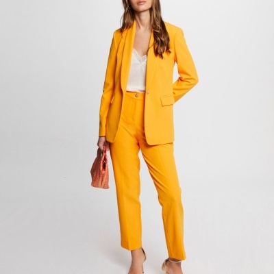 Mulher vestindo blazer e calças amarelas com blusa branca, segurando carteira cor de rosa e usando salto dourado