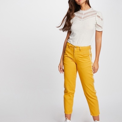 Mulher com calças mostarda até ao tornozelo, t-shirt branca com renda, ténis brancos, fundo neutro