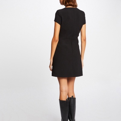 Vestido preto curto de manga curta visto de costas, com botas pretas até ao joelho, fundo claro neutro