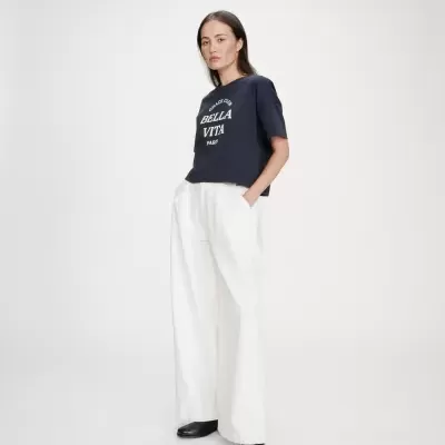 Mulher com camiseta azul escura com texto branco, calças brancas largas e sapatos pretos em fundo branco.