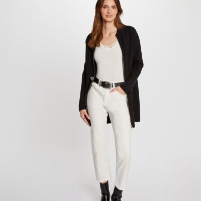 Mulher com casaco preto aberto, top branco, calças brancas com cinto preto e botas pretas, fundo branco