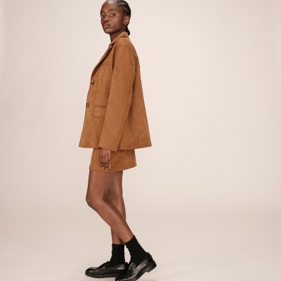 Mulher de perfil com blazer castanho, saia curta castanha, meias pretas e sapatos pretos