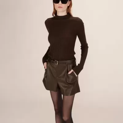 Mulher a usar camisola de malha castanha escura, calções de pele castanhos, collants pretos e óculos de sol pretos.