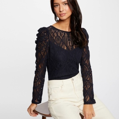 Modelo com blusa preta de malha rendada e calças claras sentada num banco de madeira