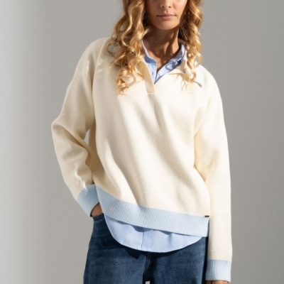 Mulher com camisola creme e azul claro sobre camisa azul, calças de ganga azuis, fundo branco.